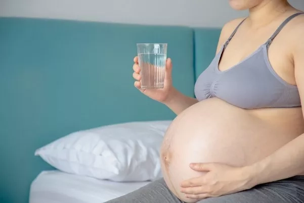 Pregnant woman sitting down on the bed with a glass of water