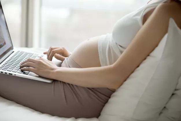 Pregnant mom sitting down on the bed and using her laptop.